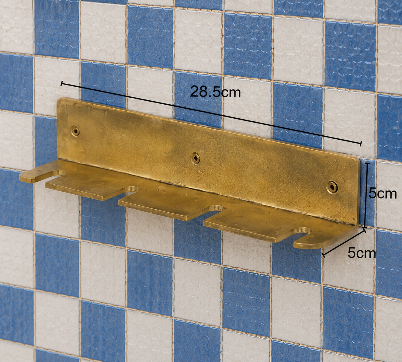 A brass wall-mounted shelf bracket with three horizontal shelves, shown against a blue and white checkered tiled wall with dimension lines indicating 28.5cm width, 5cm depth, and 5cm height.