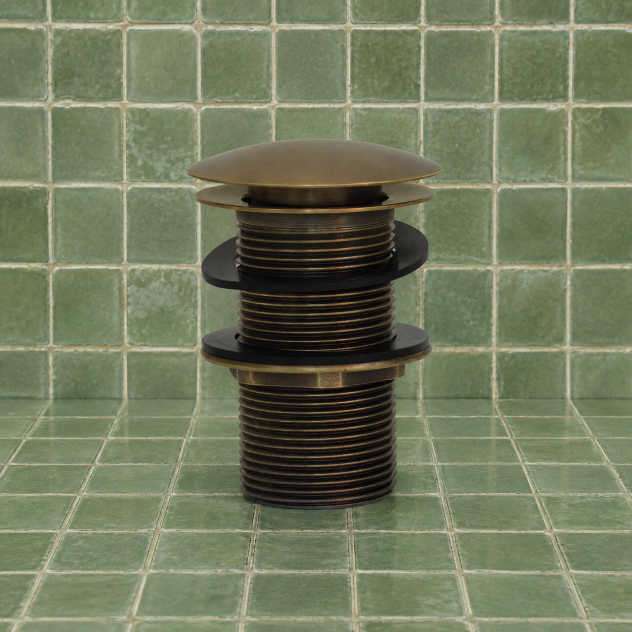 A brass kitchen sink pop-up drain assembly with black rubber gaskets, set against a green tiled background.