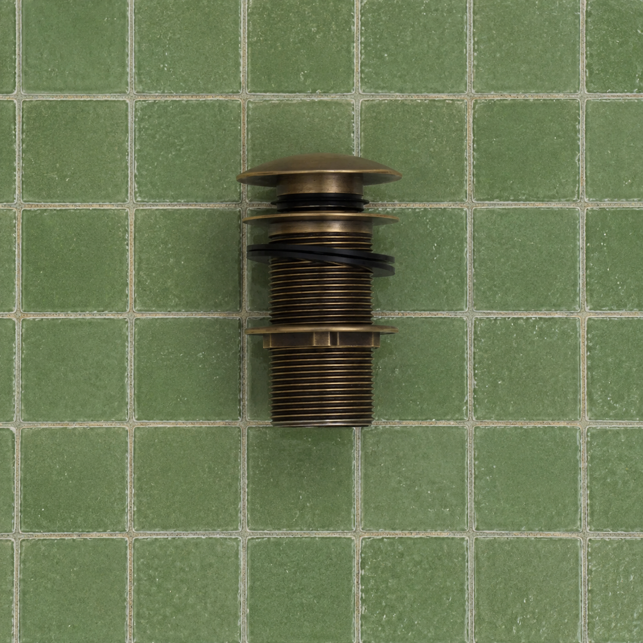 A bronze-colored metal plumbing fixture component, likely a drain stopper or valve assembly, shown against a background of green square ceramic tiles.