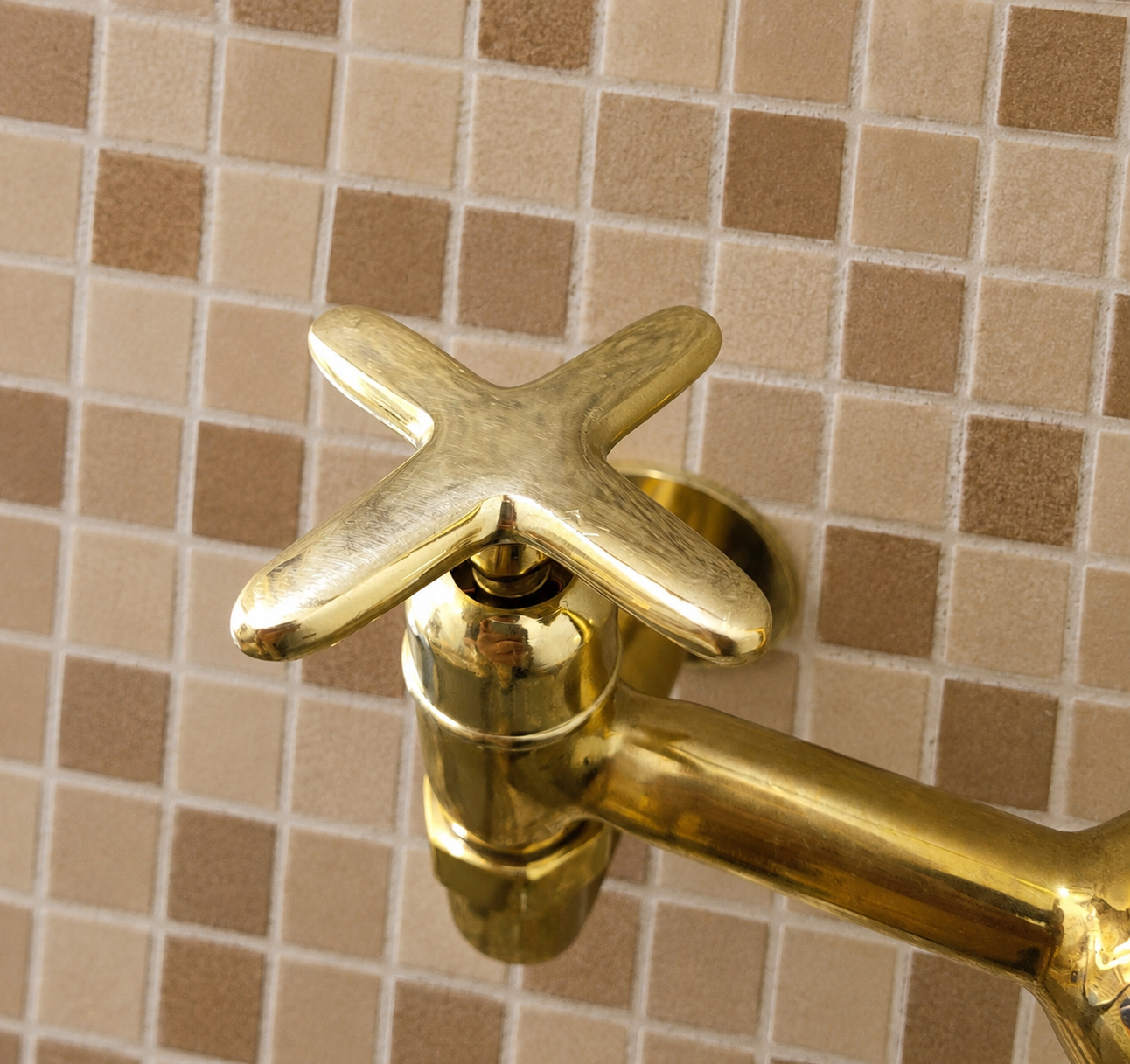 A close-up of a polished brass cross-handle faucet mounted against a beige and brown mosaic tile wall.