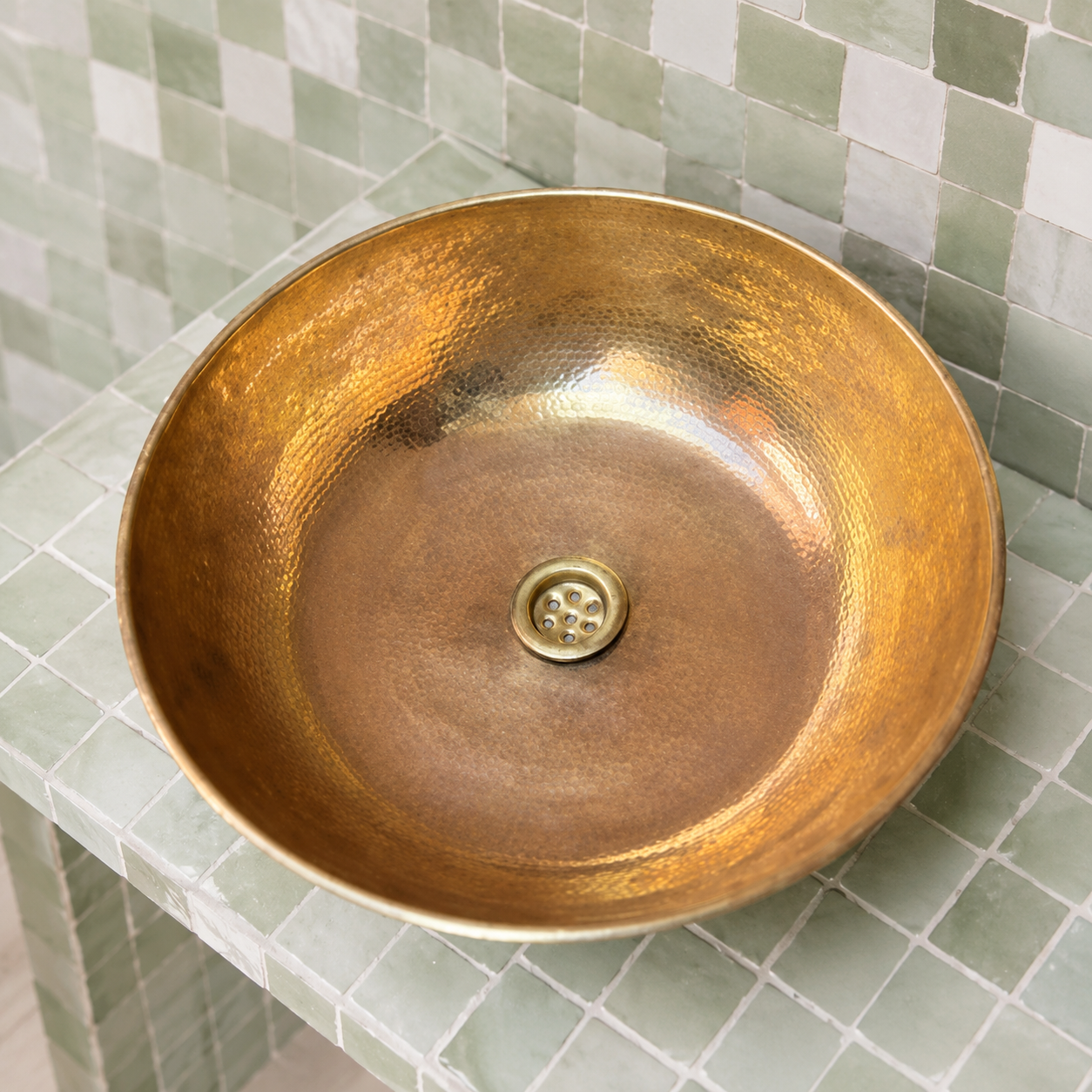 A hammered brass bathroom vessel sink basin with a matching drain, shown installed on a light green tiled countertop against a matching tiled backsplash.