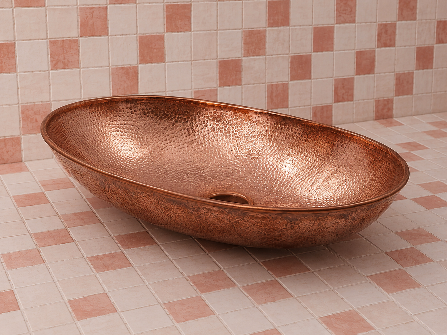 A hammered copper oval vessel sink basin with a drain, shown on a pink and white checkered tiled floor and matching tiled wall.