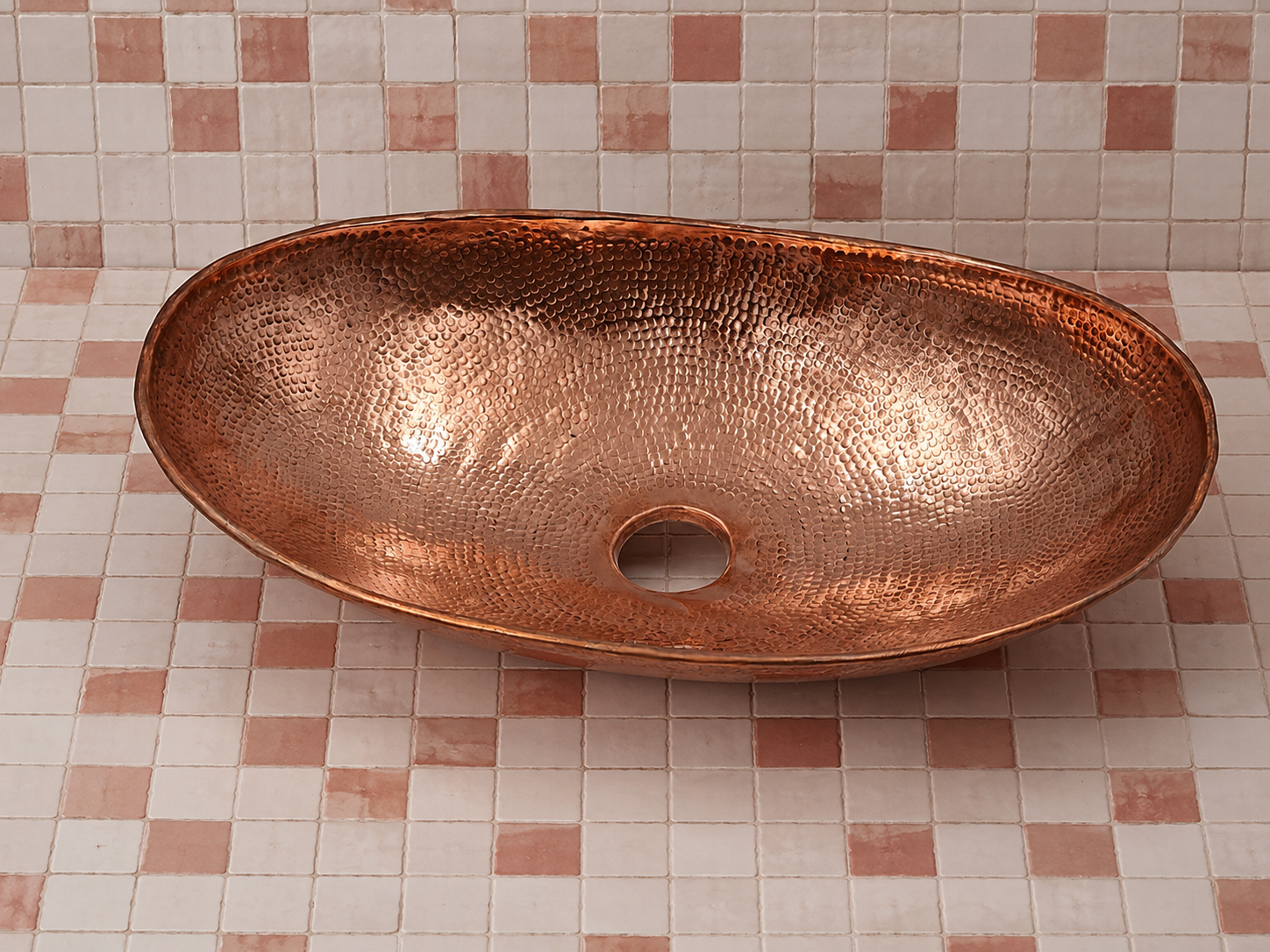 A hammered copper vessel sink basin with a central drain hole, displayed on a pink and white checkered tile surface against a matching tiled wall.