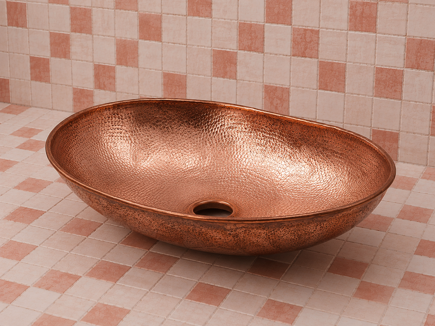 A hammered copper vessel sink basin with a matching drain, shown on a pink and beige checkered tiled floor and wall.