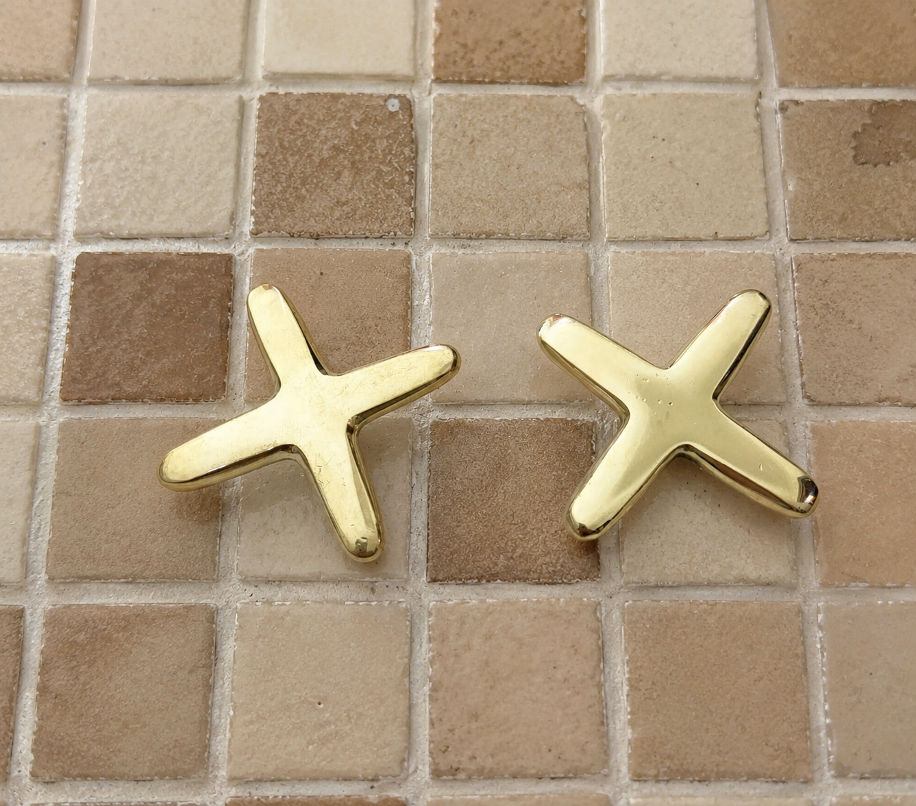 A pair of gold star-shaped stud earrings resting on a beige and brown mosaic tile surface.