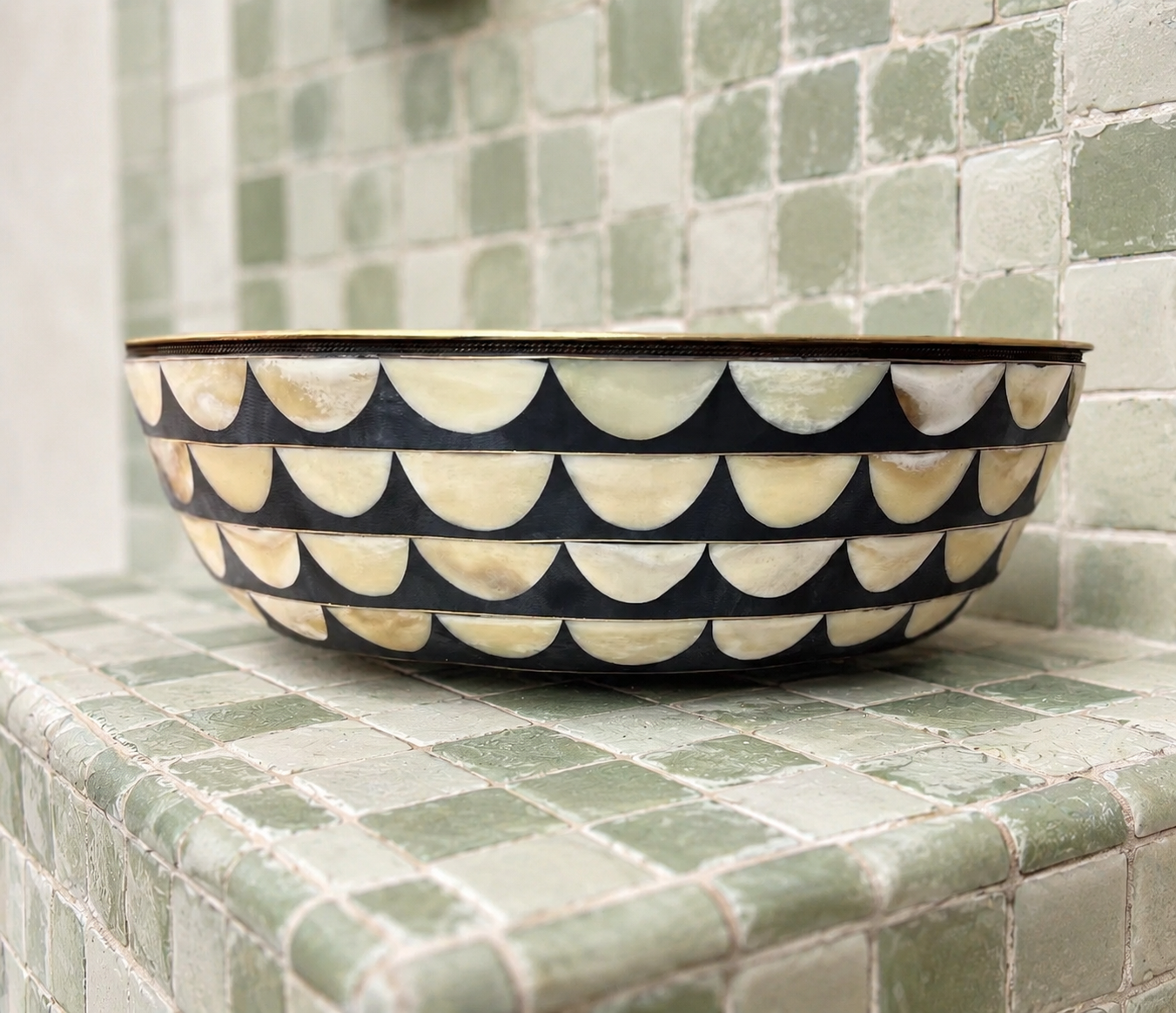 A round decorative bowl with a black and cream scalloped pattern, resting on a green tiled surface against a matching tiled background.