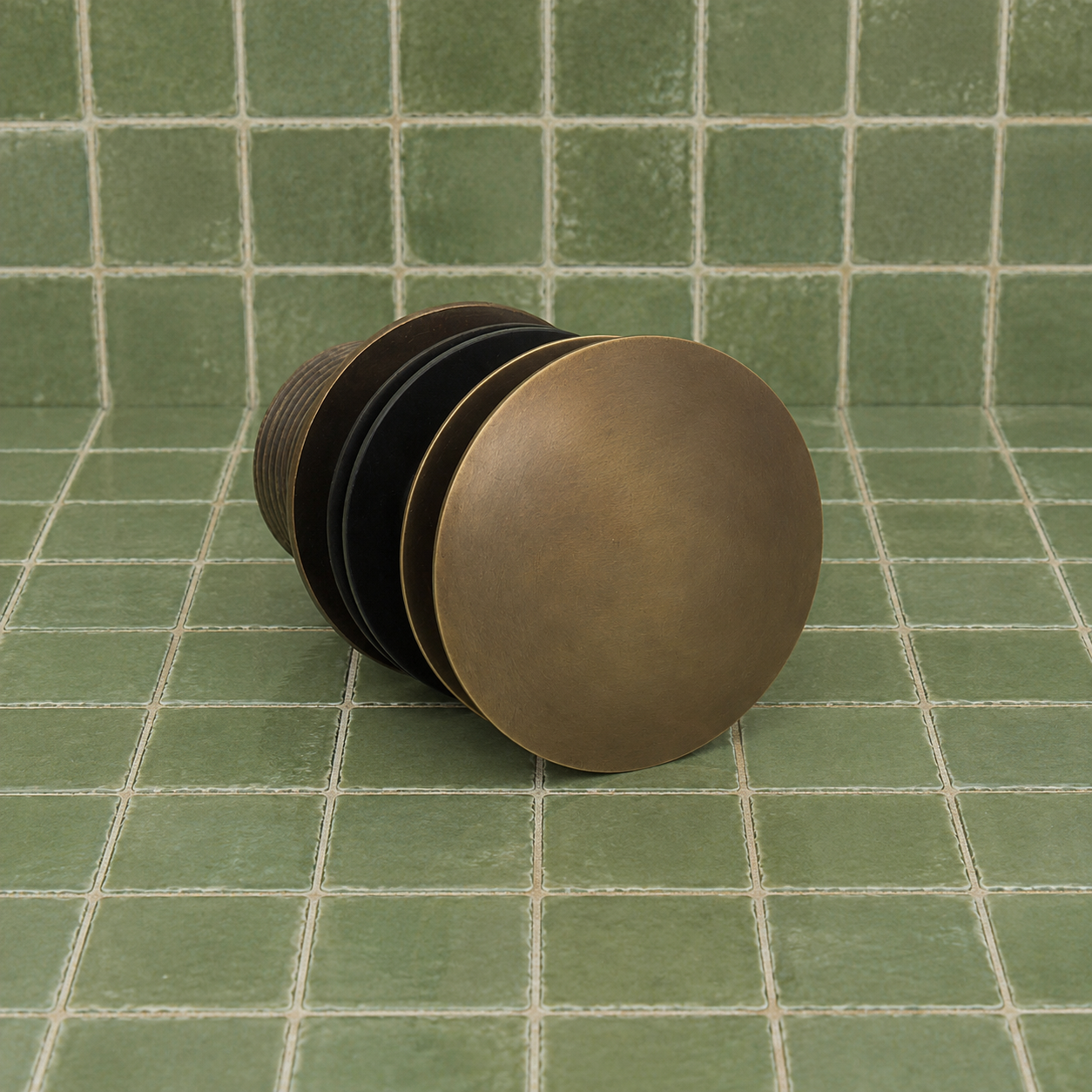 A single round metal drain stopper with a textured bronze finish, featuring a solid top dome and ridged sides, resting on a green tiled surface.