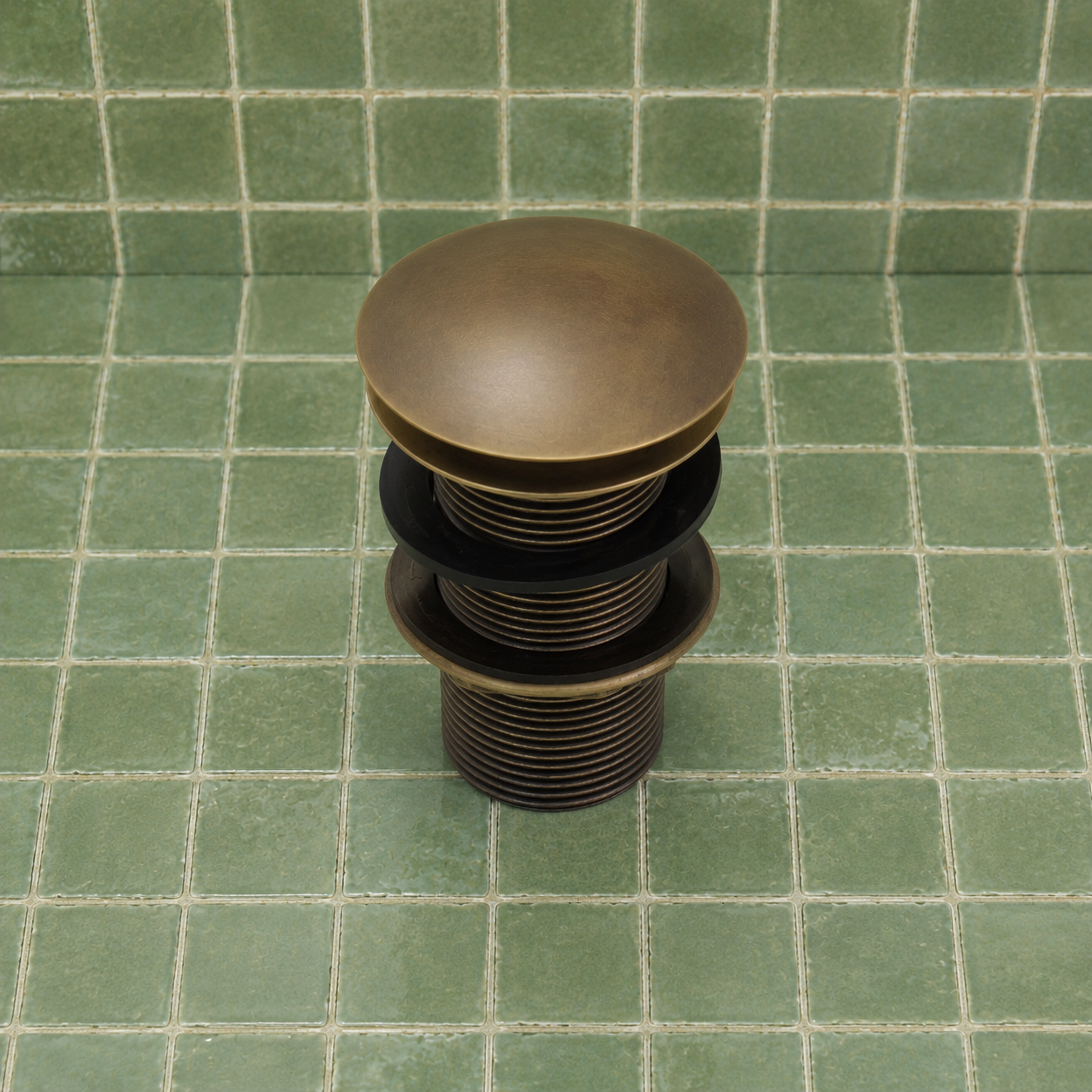 A single round metal drain stopper with a textured bronze finish, featuring a solid top dome and ridged sides, resting on a green tiled surface.
