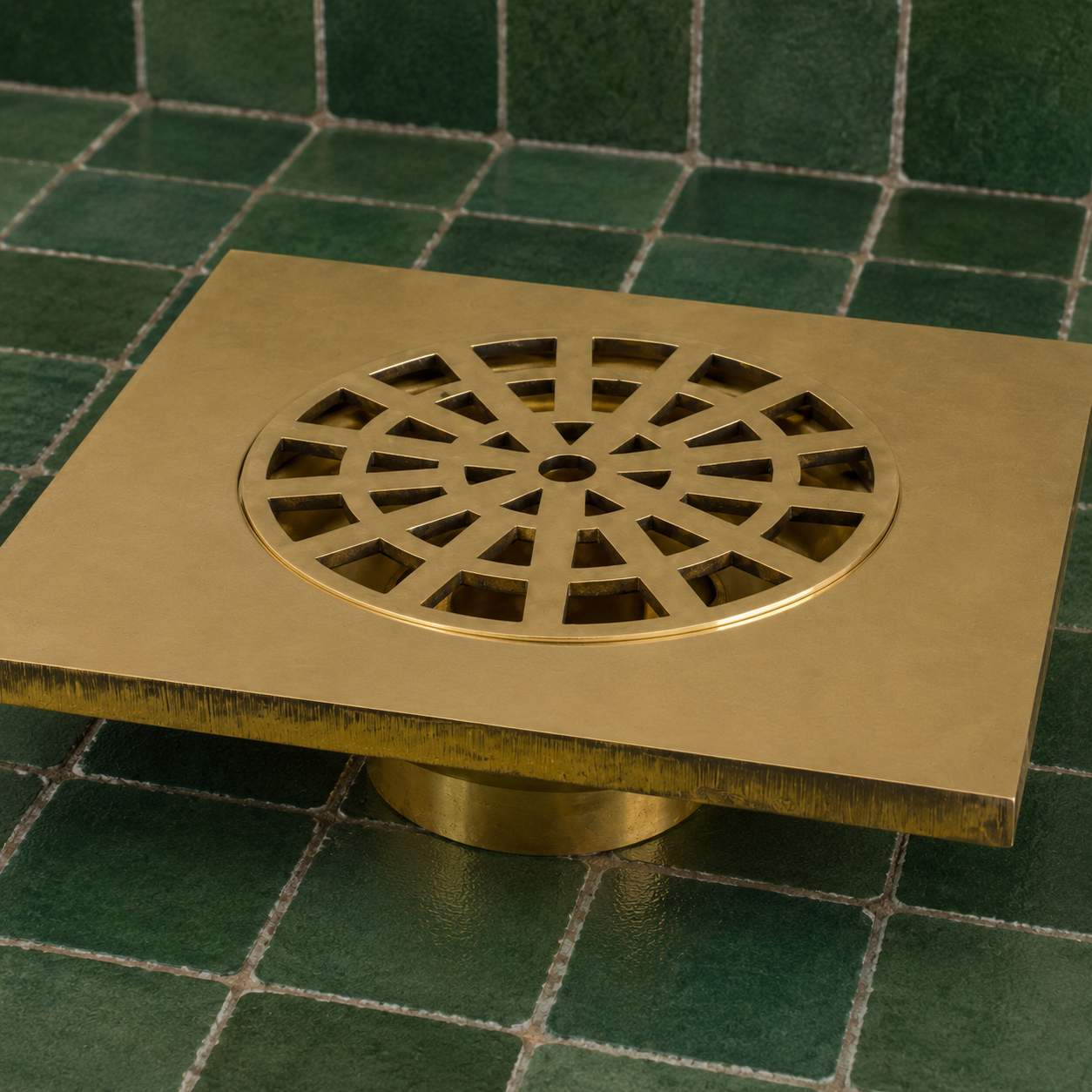 A square brass floor drain with a circular cutout and decorative radial pattern on top, set against a green tiled background.