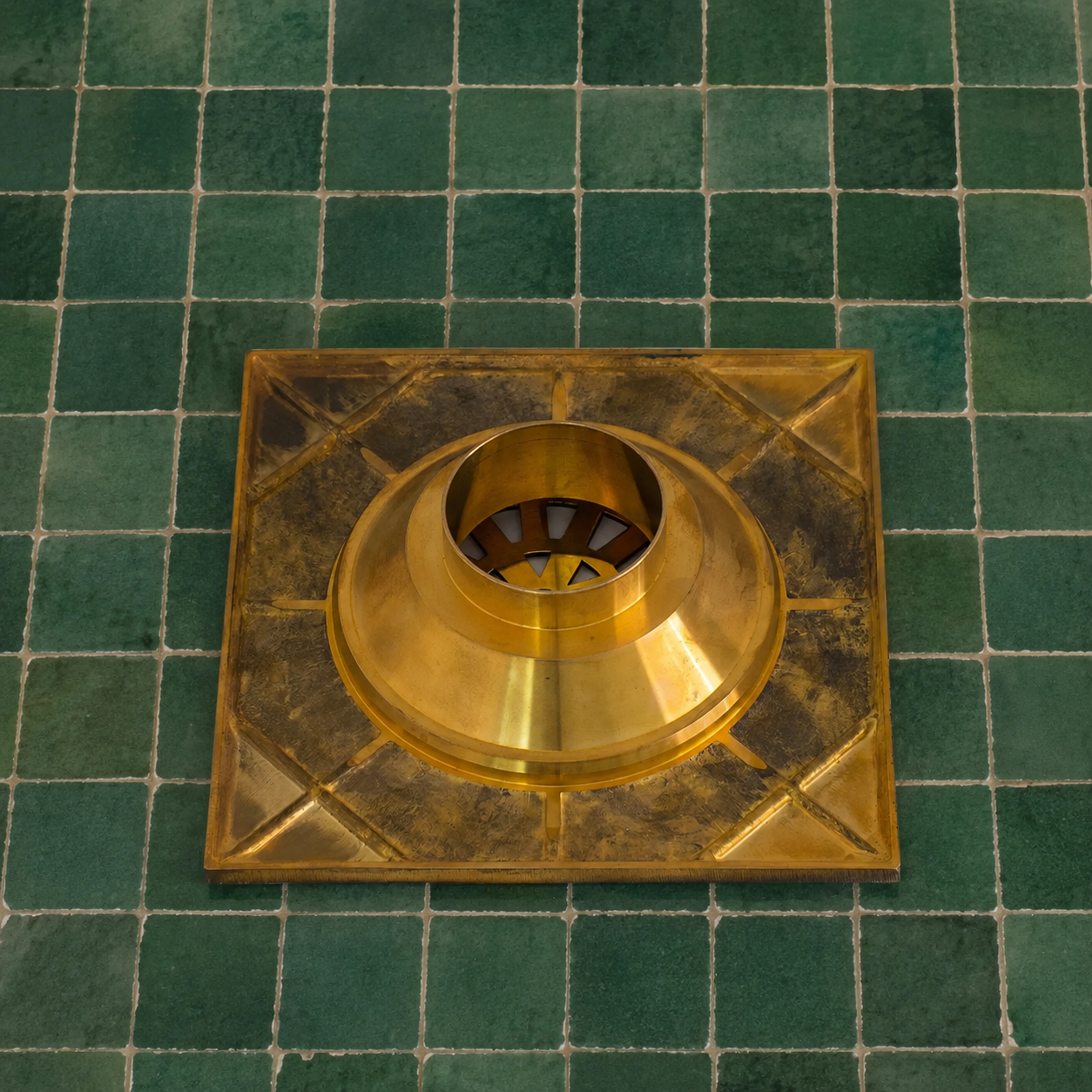 A square brass metal sink drain cover with a central round drain opening, placed on a green tiled surface.