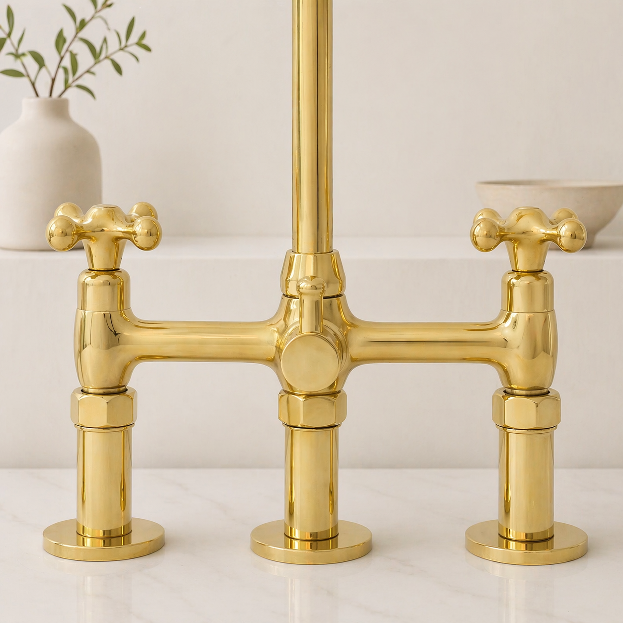 A polished gold bridge faucet with three cross-shaped handles, shown on a white marble countertop with a neutral background and a small white vase.