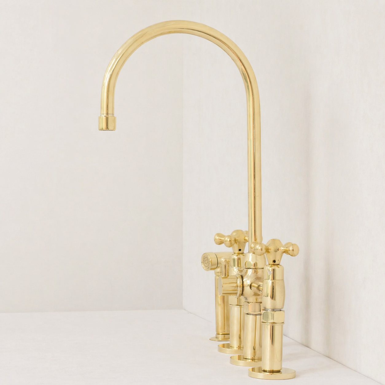 A polished brass kitchen faucet with a high-arc spout and two separate cross-handle valves, shown installed against a white wall and white countertop.