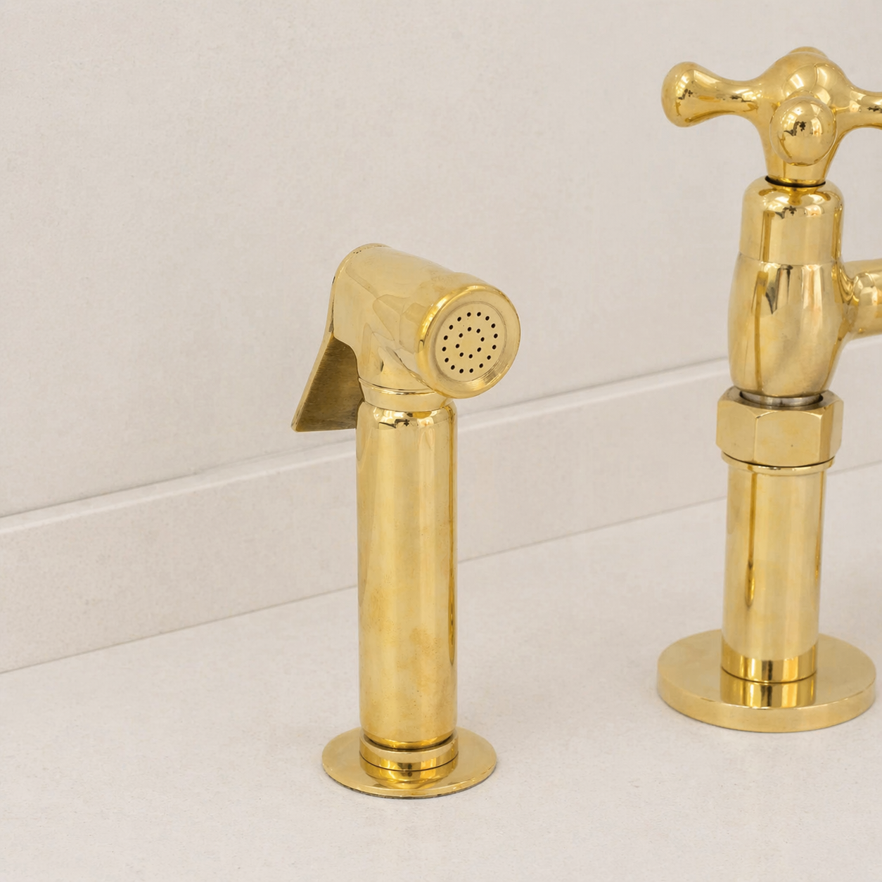 Gold-finished bathroom faucet set with a side sprayer and cross-handle faucet on a light beige countertop against a white tiled wall.