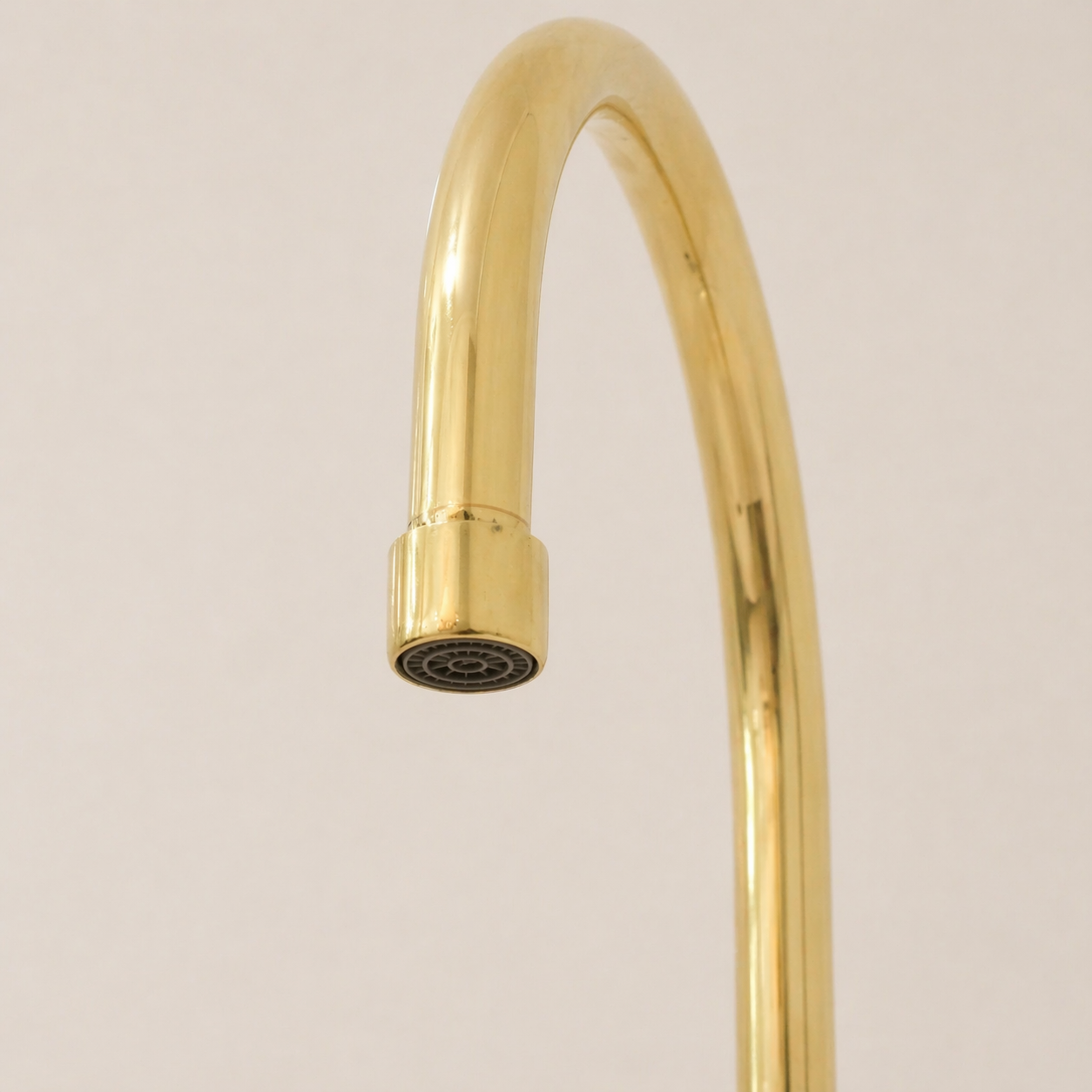 A close-up of a polished gold faucet with a curved spout and a round spray head, shown against a plain light beige background.