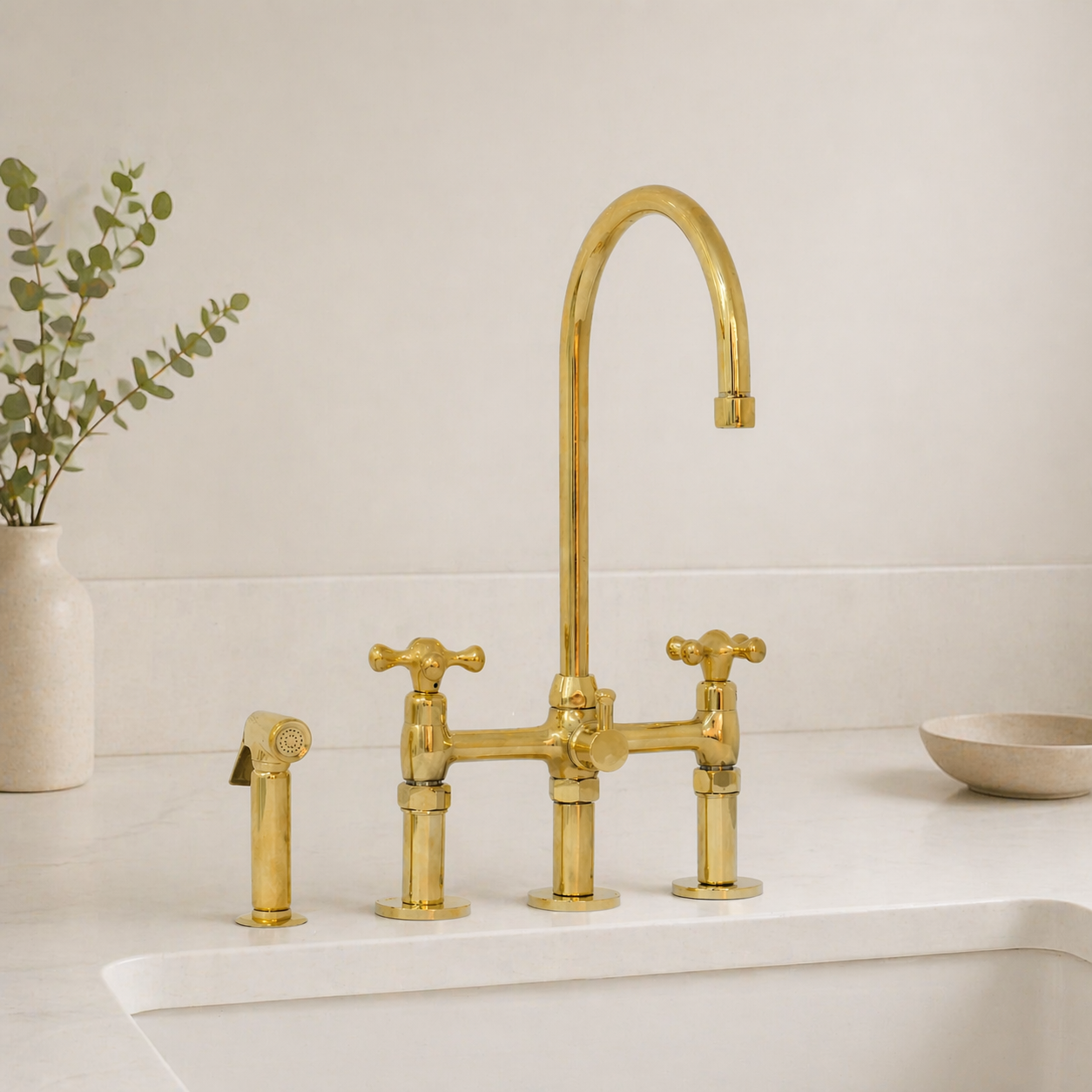 A polished brass three-hole bridge faucet with a high-arc spout, two cross-handle valves, and a matching side sprayer, installed on a white sink with a warm cream background and a small vase of eucalyptus.