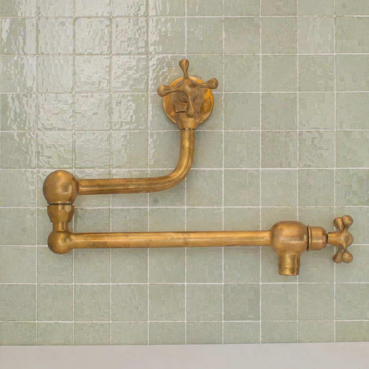 Handmade unlacquered solid brass wall-mounted pot filler faucet with double swing arms and dual shut-off valves installed above a kitchen stove.