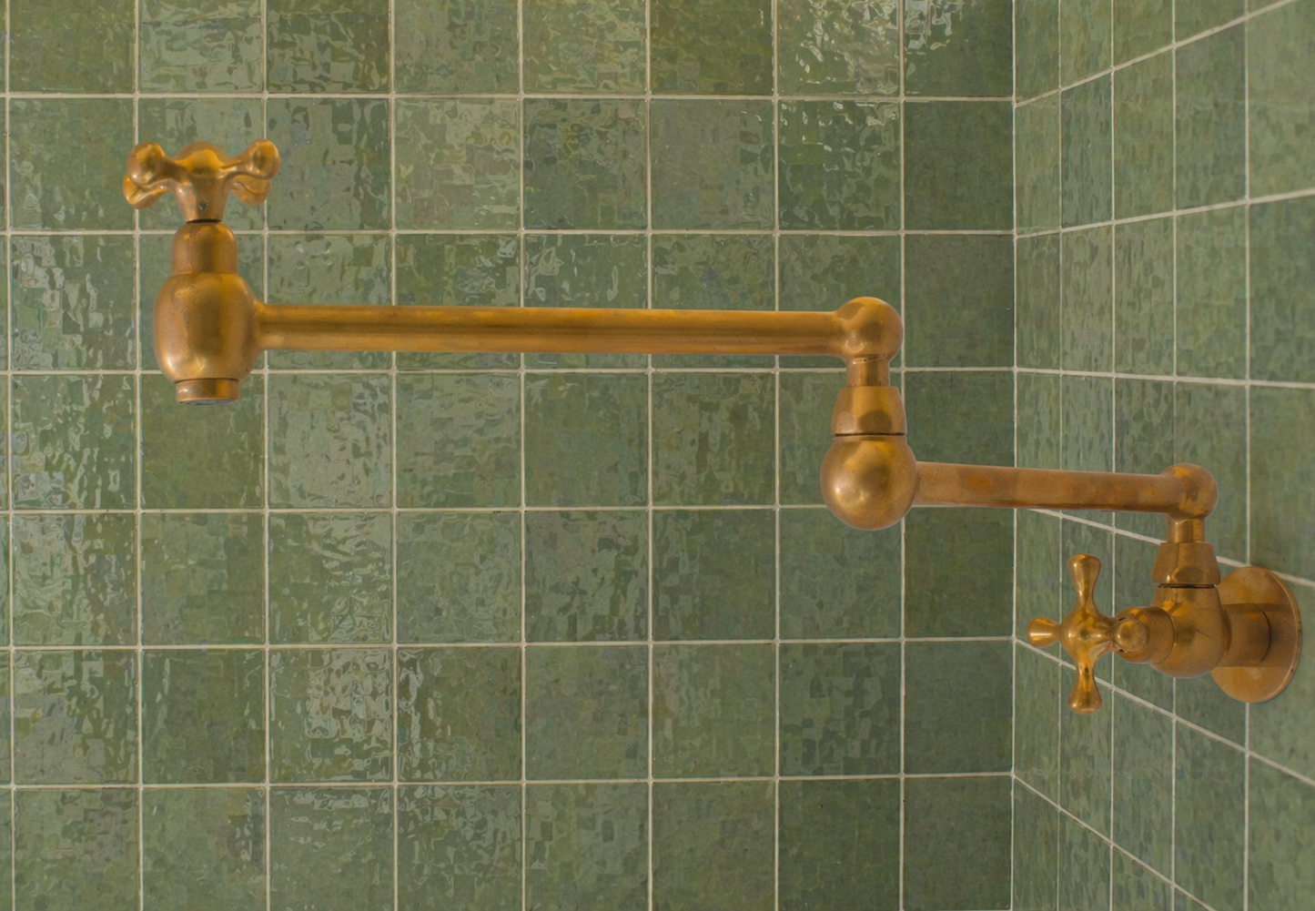 Handmade unlacquered solid brass wall-mounted pot filler faucet with dual swing arms and cross handles installed above a kitchen stove.