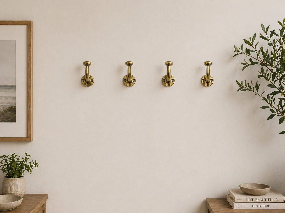 Four brass wall hooks mounted in a row on a warm off-white wall, styled in a minimalist interior with a framed print, small potted plants, and books.