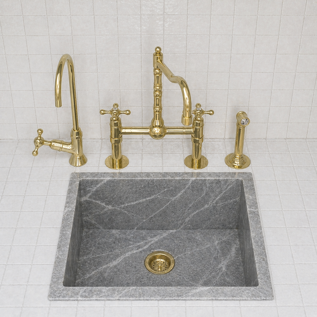 A modern kitchen sink setup featuring a square gray stone basin with a brass drain, a polished brass bridge faucet with a high-arc spout, a separate tall gooseneck faucet, and a matching side lever handle, all set against a white tiled backsplash.