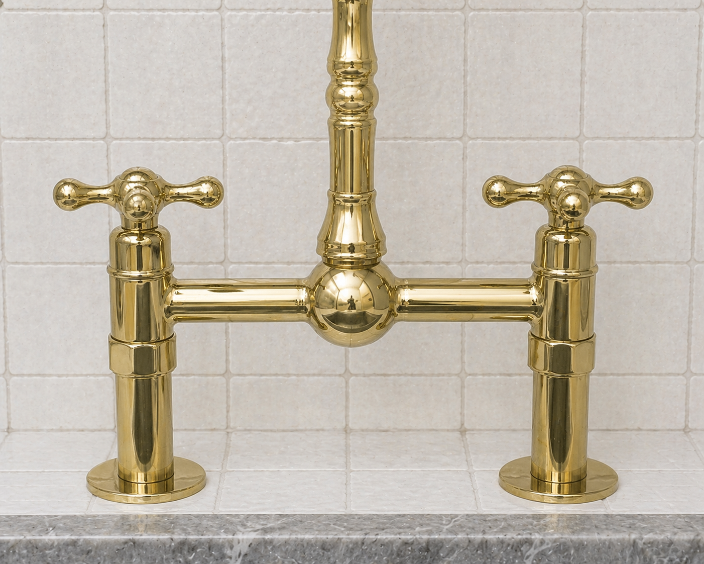 A polished brass bridge faucet with two cross handles, mounted on a gray marble countertop against a white tiled backsplash.