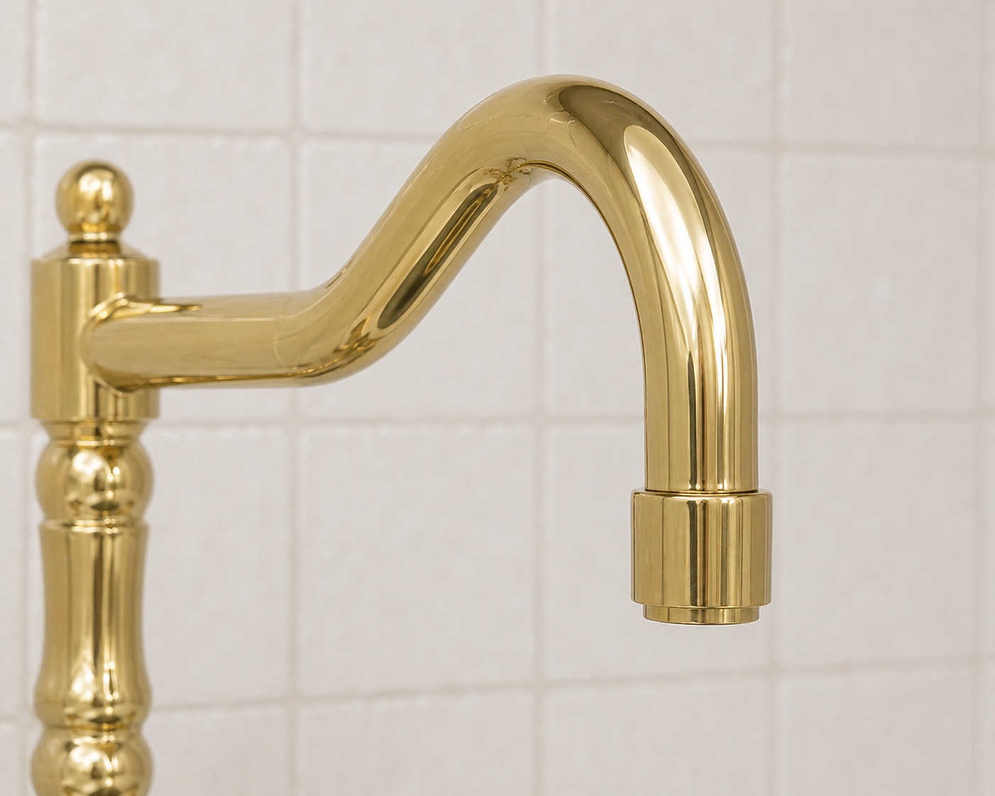 A polished gold brass kitchen faucet with a curved spout and a turned decorative column base, shown against a white tiled wall.