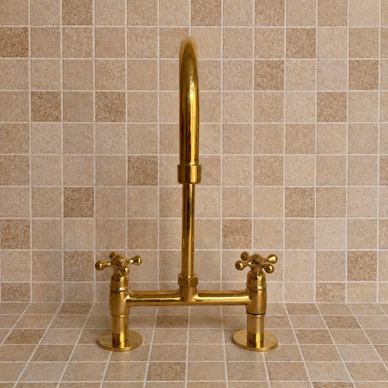 A polished brass wall-mounted kitchen faucet with a high-arc spout and two cross-handle valves, installed against a beige tiled backsplash.