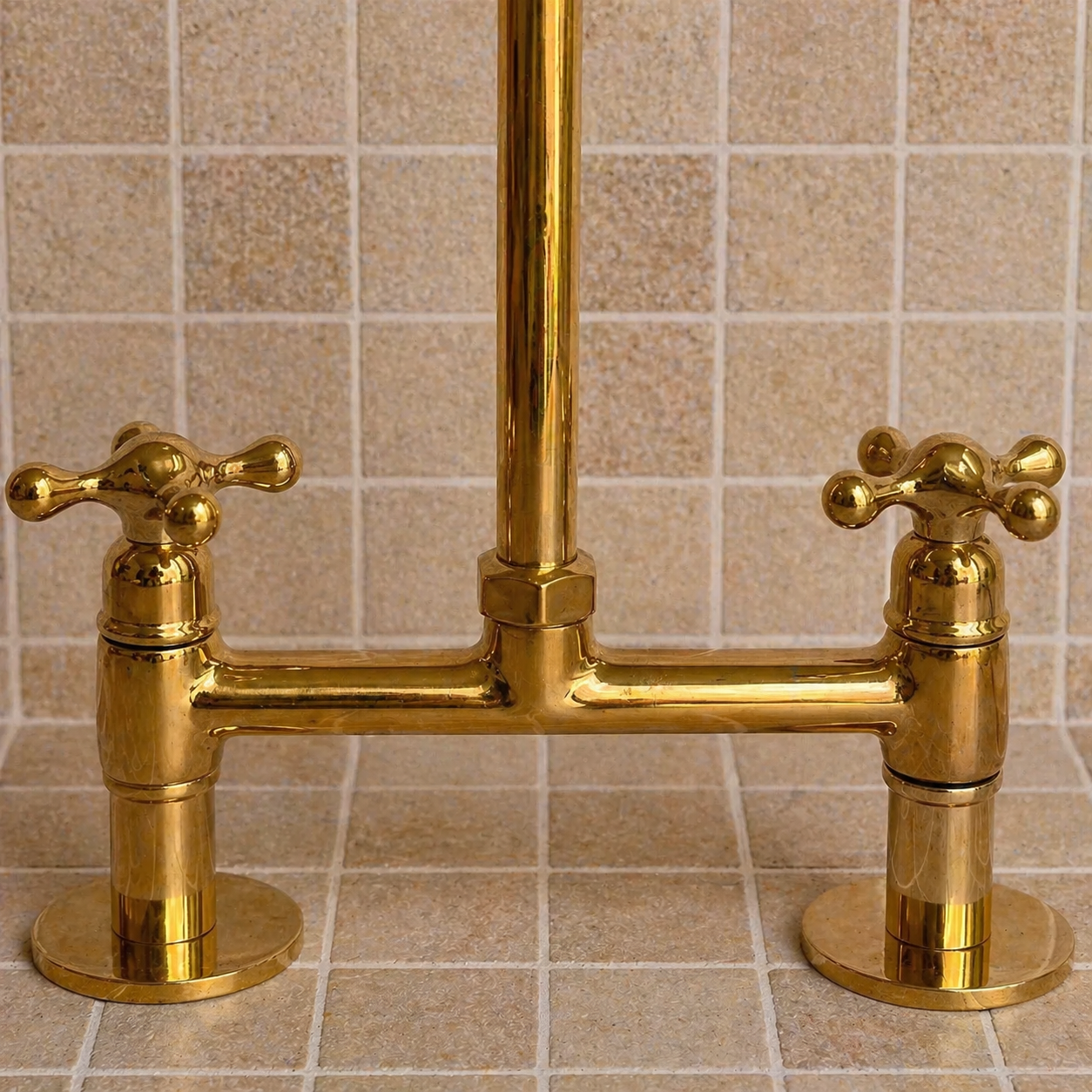 A polished brass two-handle bridge faucet set mounted against a beige tiled wall and countertop.