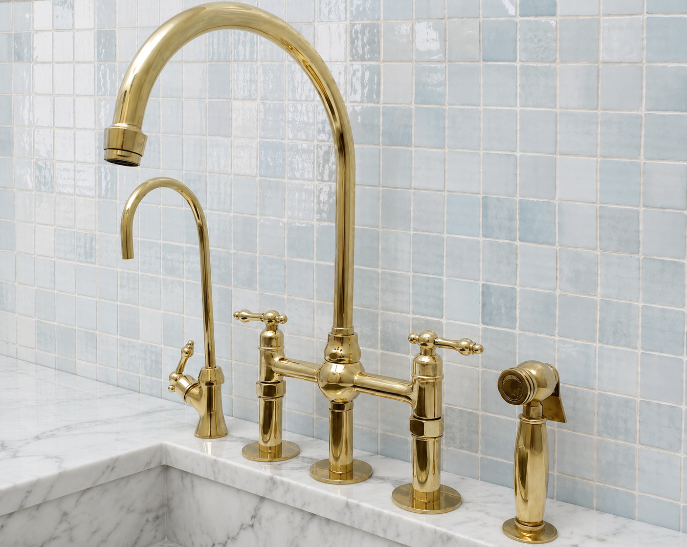 A polished gold bridge faucet set installed on a white marble sink, with a matching high-arc spout, a smaller side spout, and a handheld sprayer, set against a light blue mosaic tile backsplash.