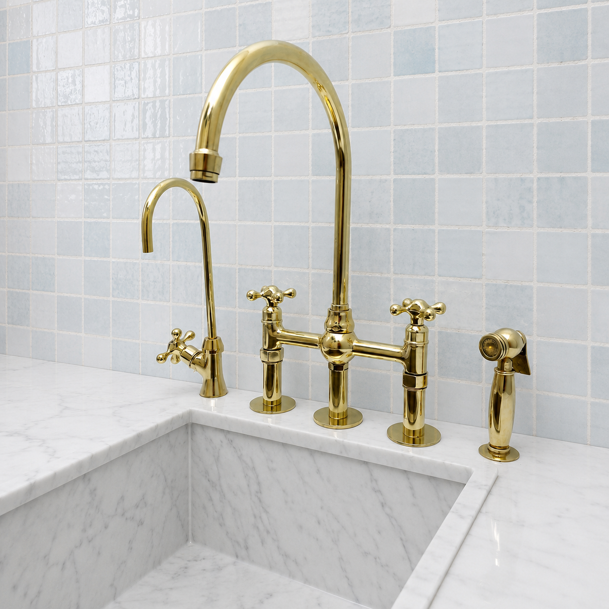 A polished gold bridge faucet set installed above a white marble sink, with a light blue tiled backsplash behind it. The set includes a tall gooseneck main faucet, a smaller auxiliary faucet, a side sprayer, and two cross handles.