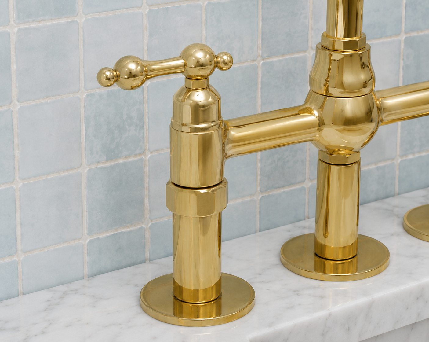 A close-up of a polished gold double-handle bathroom faucet mounted on a white marble countertop against a light blue tiled backsplash.