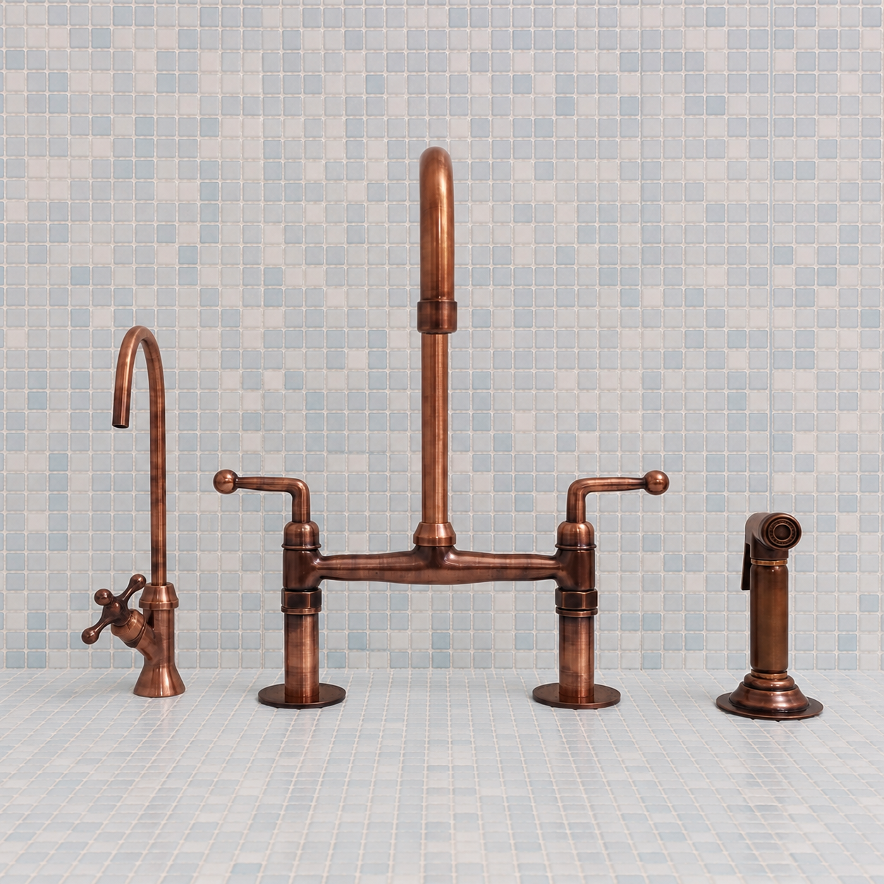 A copper bridge faucet set with a tall gooseneck spout, a separate hot and cold supply valve, and a matching side sprayer, displayed against a light blue mosaic tile backsplash.