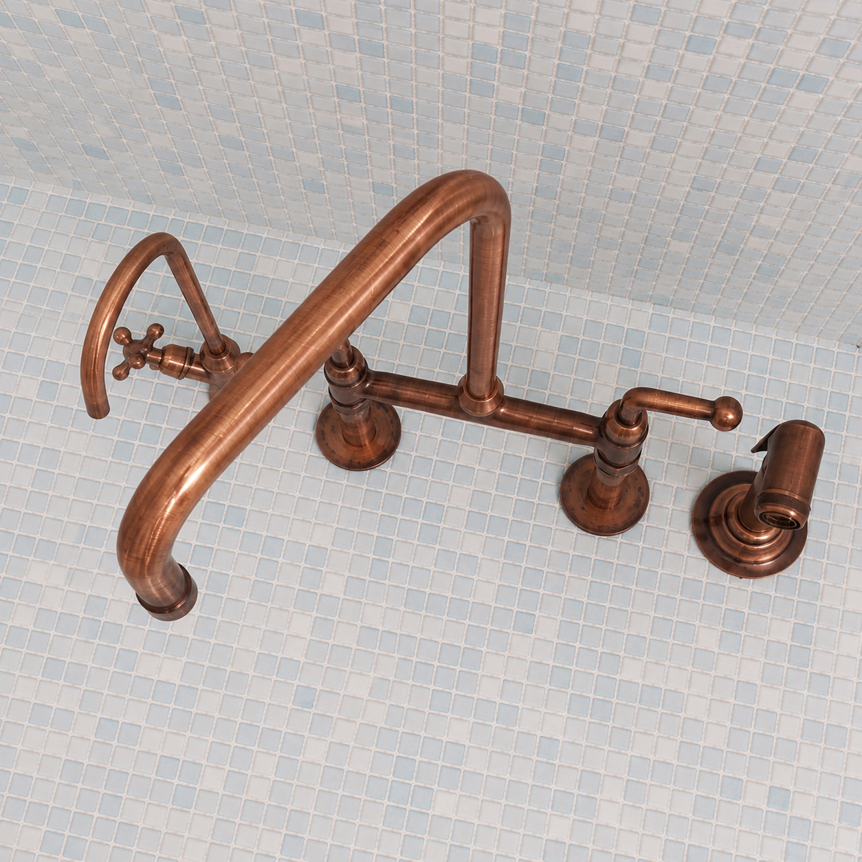 A copper bridge faucet set with a curved main spout, two separate handles, and a matching drain cover, shown against a light blue mosaic tile background.