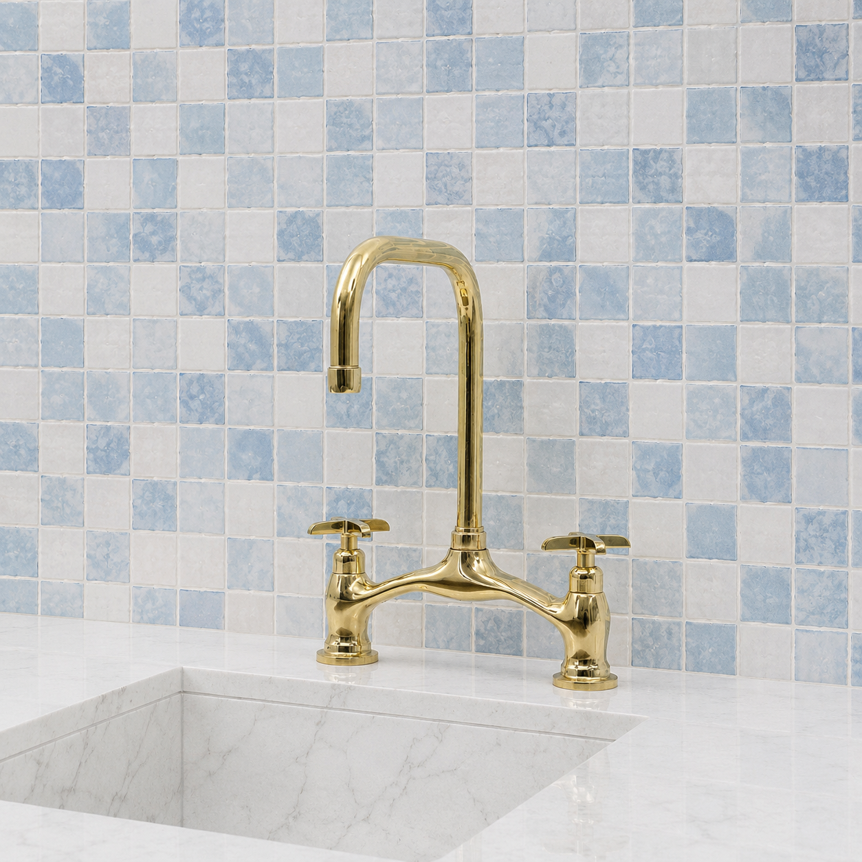 A polished gold bridge faucet with two separate handles, installed on a white marble sink against a blue and white checkered tile backsplash.