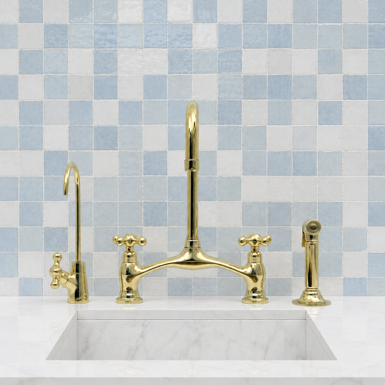 A polished gold three-hole kitchen faucet set with a tall central spout, two cross-handle cold faucets, a side lever, and a handheld sprayer, installed over a white marble sink against a light blue mosaic tile backsplash.