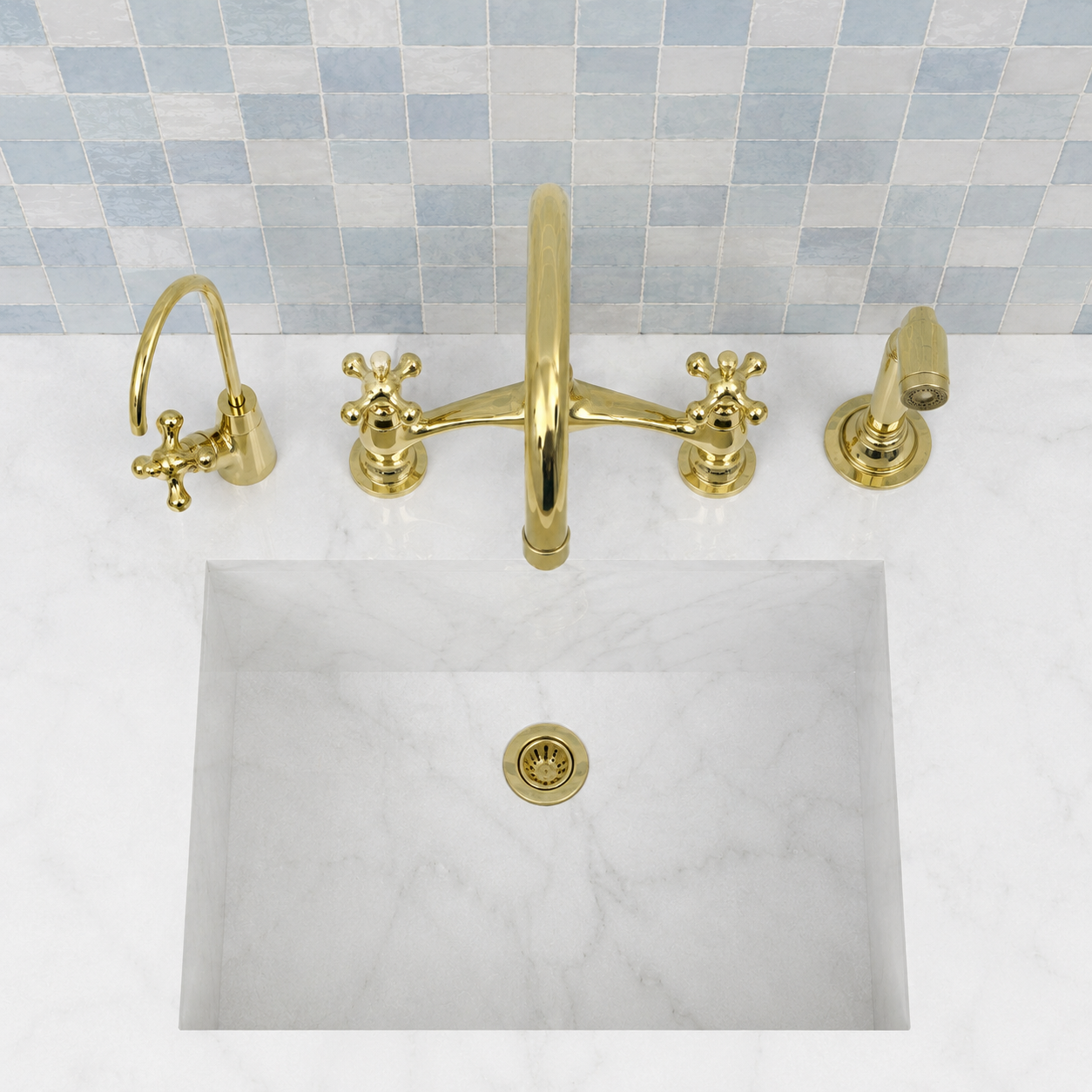 A polished gold brass faucet set with a high-arc central spout, two cross-handle faucets, and a separate wall-mounted spray head, installed above a white marble sink with a light blue mosaic tile backsplash.