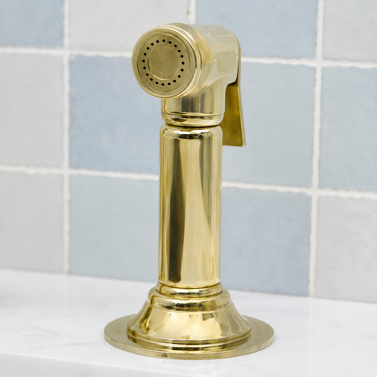 A polished brass kitchen sink sprayer with a circular shower head and side lever, mounted on a white sink countertop in front of a light blue tiled wall.