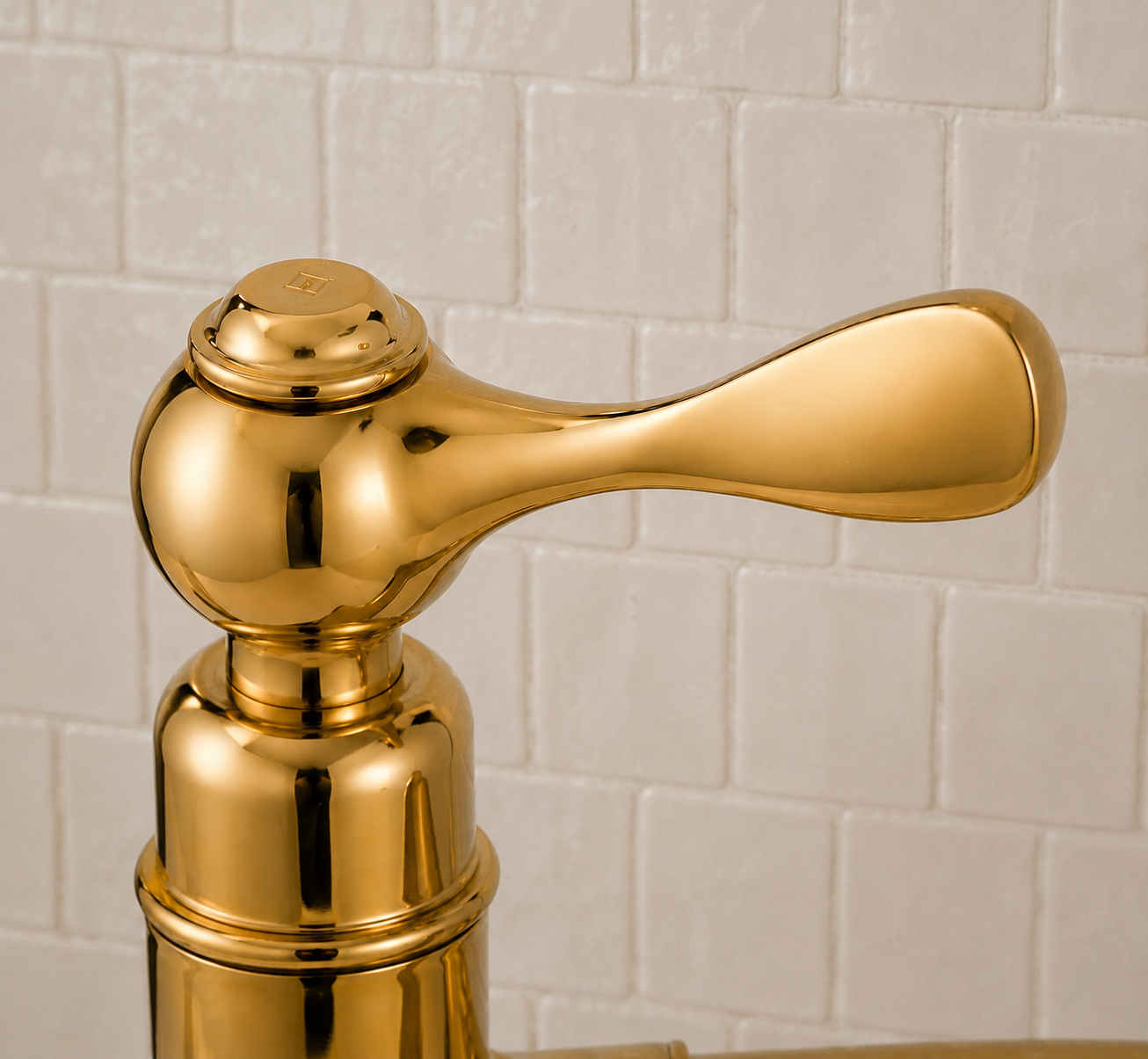 A close-up of a polished gold bathroom faucet handle with a matching cylindrical base, set against a light beige tiled wall background.