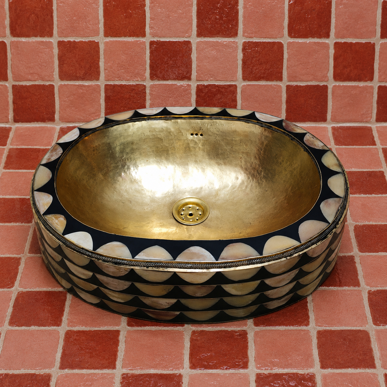A round vessel sink with a hammered gold-toned interior and a black exterior decorated with scalloped mother-of-pearl shell patterns, set against a red tiled wall.