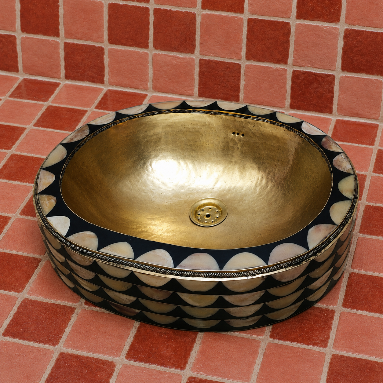 An oval vessel sink with a hammered gold interior and a black exterior decorated with a scalloped shell pattern, set against a red tiled background.