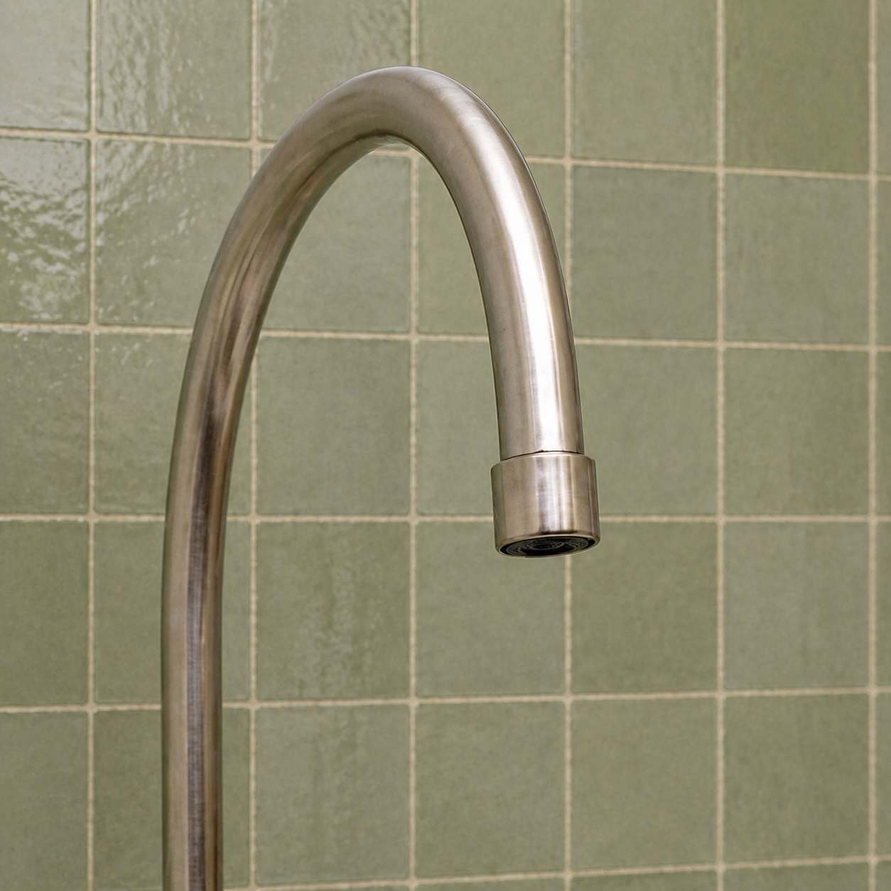 A curved brushed nickel kitchen faucet shown against a background of green square ceramic tiles.