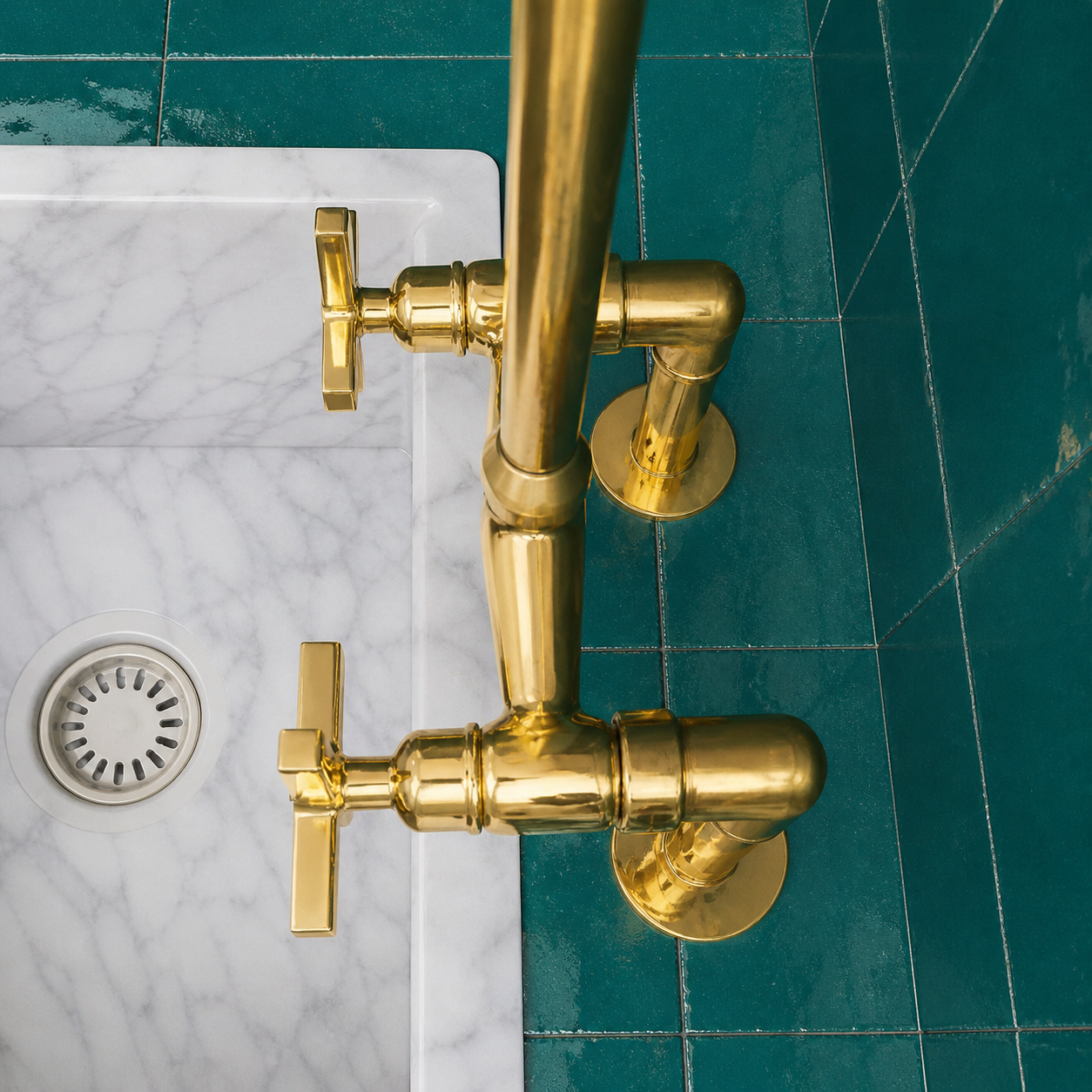 A close-up of a polished brass wall-mounted faucet with two cross handles, installed above a white marble sink on a dark teal tiled wall.