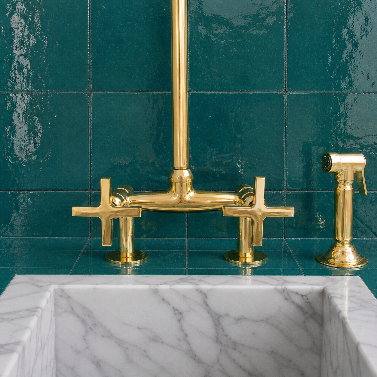 A polished brass bridge faucet set with two cross-shaped handles and a matching side sprayer, mounted against a glossy teal tiled wall above a white marble sink.