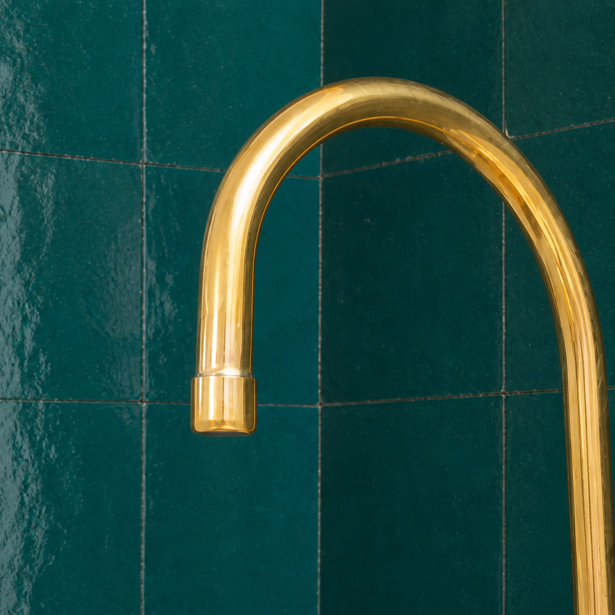 A curved, polished brass faucet set against a background of glossy dark green tiles.