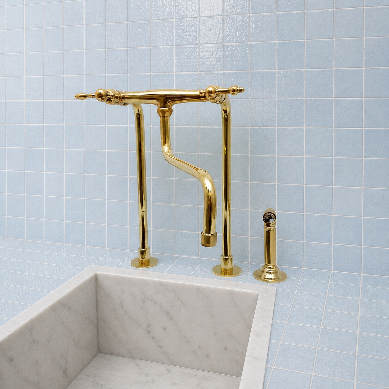 A polished gold bridge faucet set with a curved spout, two vertical handles, and a separate side sprayer, installed above a white marble sink against a light blue tiled wall.