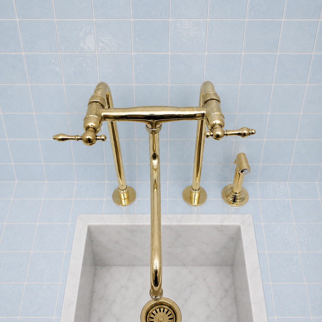 A polished brass bridge faucet with two cross handles and a matching side sprayer, mounted above a white marble sink against a light blue tiled wall.