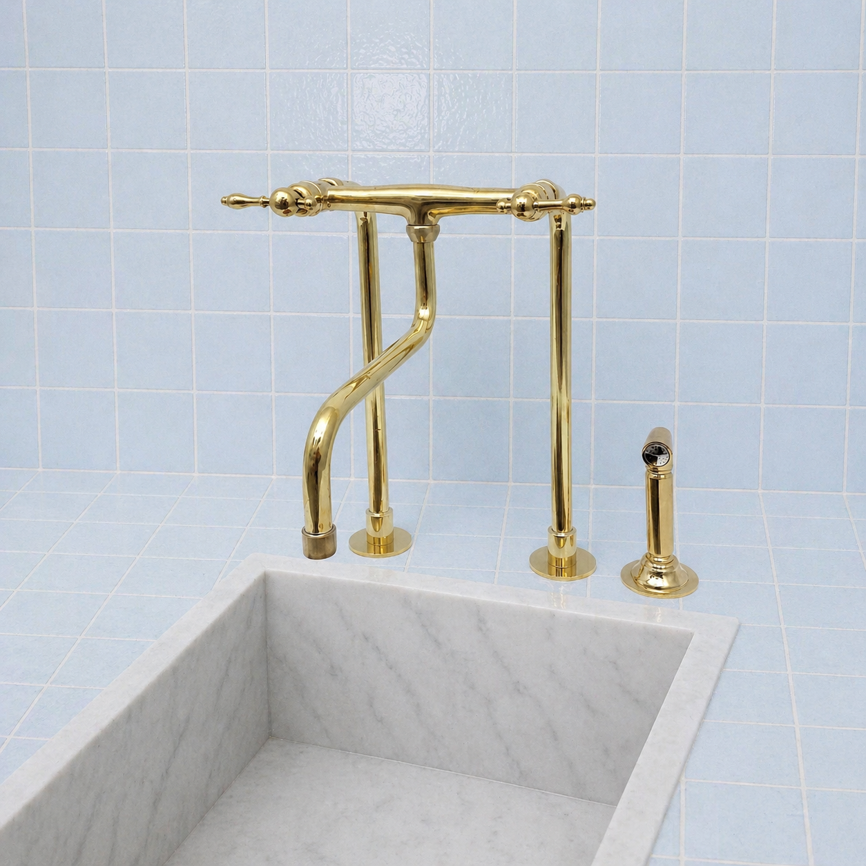 A polished gold bridge faucet set with a curved spout, two vertical handles, and a separate side sprayer, installed above a white marble sink against a light blue tiled wall.