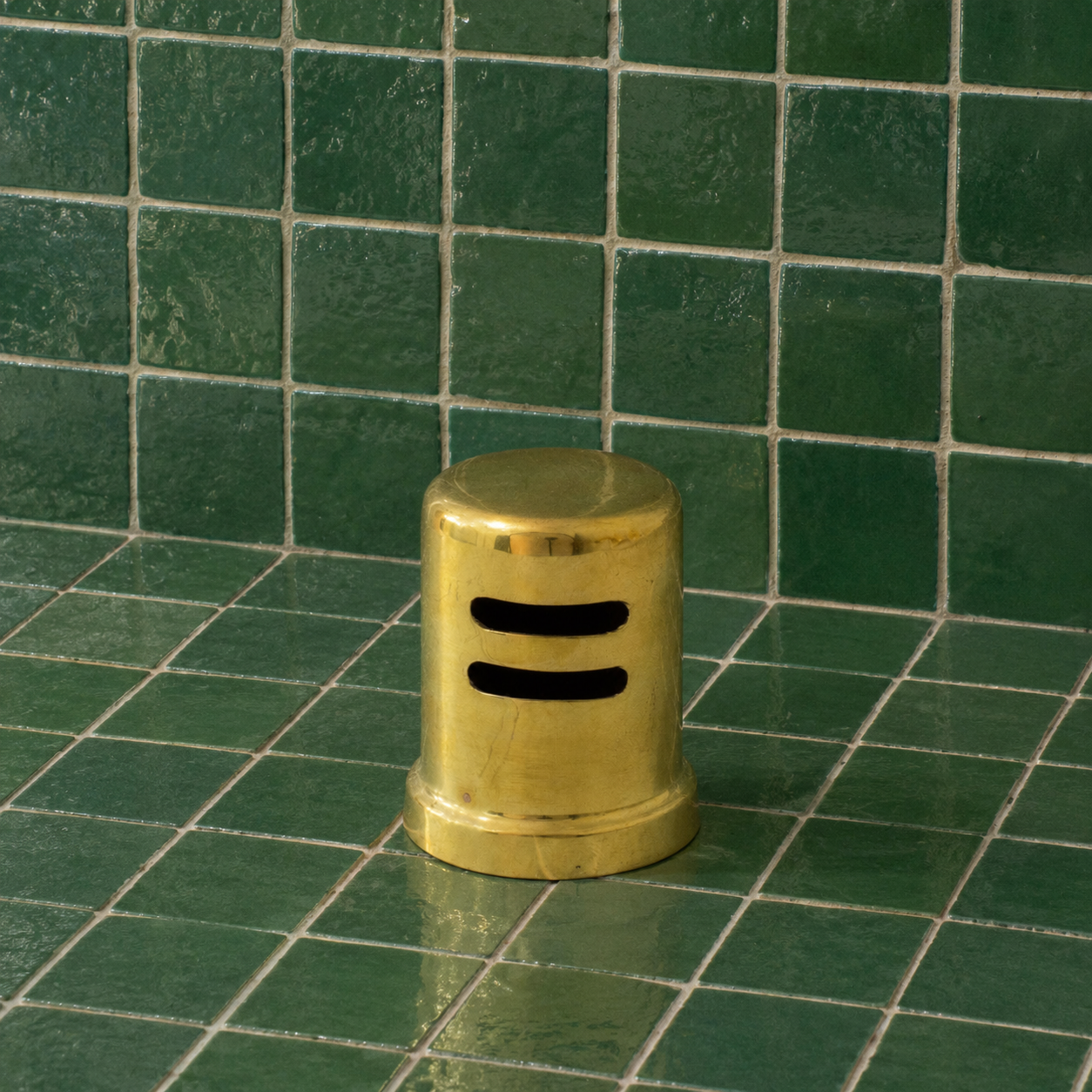 A small cylindrical brass object with two horizontal black slots on the front, standing on a glossy green tiled surface against a matching tiled wall.