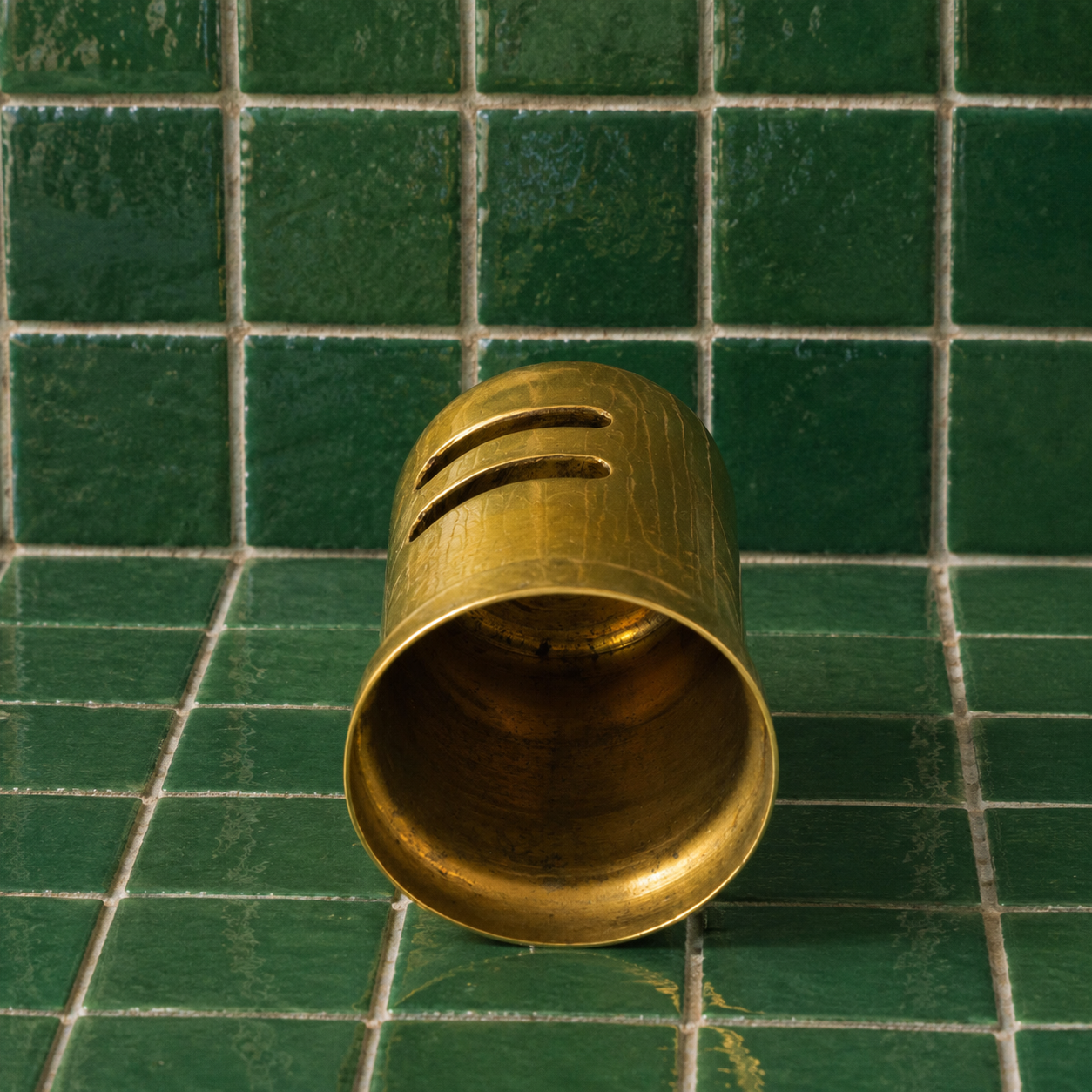 A brass bell-shaped cup with three horizontal slots on its side, resting on a green tiled surface.