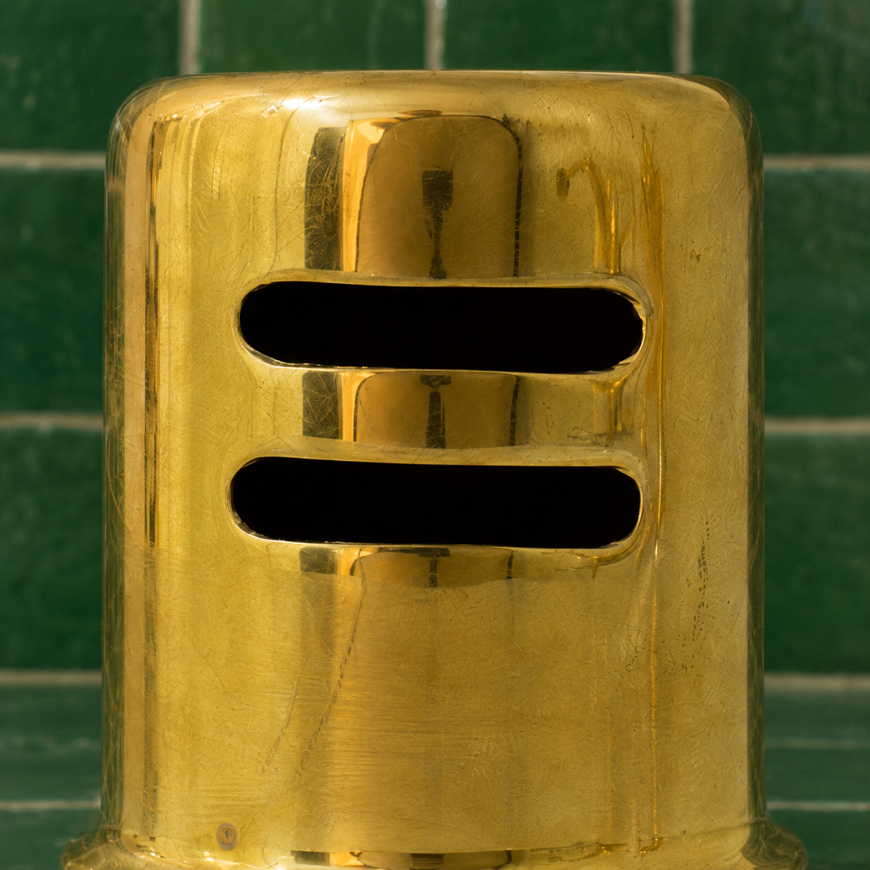 A close-up of a shiny gold metal dispenser or container with two horizontal black slots, set against a green tiled background.