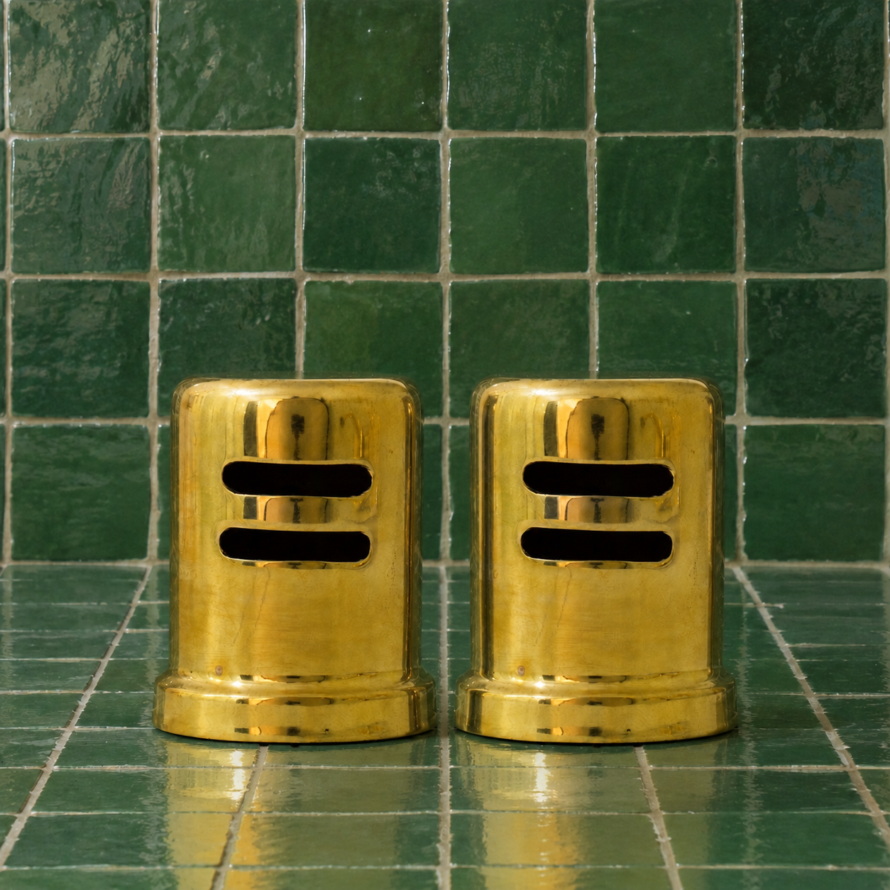 Two polished brass cylindrical salt and pepper mills with horizontal slots, standing side-by-side on a green tiled surface against a matching green tiled background.